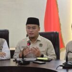 Three men in beige uniforms and black peci hats sit at a conference table during a formal briefing, with an Indonesian flag in the background and microphones in front of them.