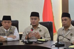Three men in beige uniforms and black peci hats sit at a conference table during a formal briefing, with an Indonesian flag in the background and microphones in front of them.