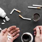 Hands near a spilling white pill bottle with several pills on a dark surface, surrounded by broken cigarettes, a glass of dark liquid, and a spiral notebook—conveying an addiction-related scene.