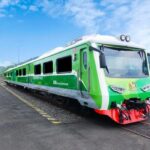 Green passenger train at a platform beside railway tracks on a sunny day with countryside in the background.