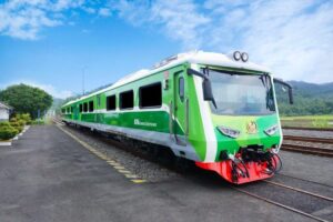 Green passenger train at a platform beside railway tracks on a sunny day with countryside in the background.