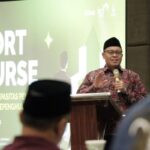 Man in batik shirt and black cap speaks at a podium during a seminar, with a large 'SHORT COURSE' banner behind.