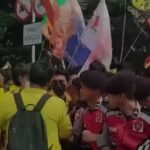 Crowd of people at an outdoor demonstration, holding colorful flags and banners with uniforms visible in the front.