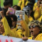 Protesters in yellow jackets raise fists, shouting slogans at a rally, holding signs and banners