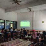 Group of students seated at tables in a classroom watching a soccer match projected on the wall.