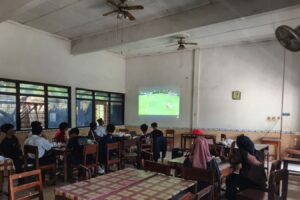 Group of students seated at tables in a classroom watching a soccer match projected on the wall.