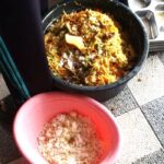 Person standing next to a large pot of mixed cooked food on a tiled floor; a pink bowl with salt sits nearby.