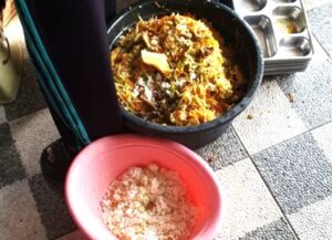 Person standing next to a large pot of mixed cooked food on a tiled floor; a pink bowl with salt sits nearby.