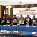 Indonesian officials stand behind a blue-draped table, displaying seized documents and evidence at a press conference with a large banner in the background.