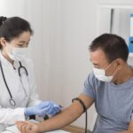Female doctor in white coat and mask administers a injection to a male patient wearing a mask in a clinic. A stethoscope hangs from her neck.