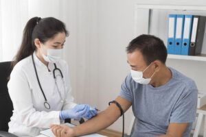 Female doctor in white coat and mask administers a injection to a male patient wearing a mask in a clinic. A stethoscope hangs from her neck.