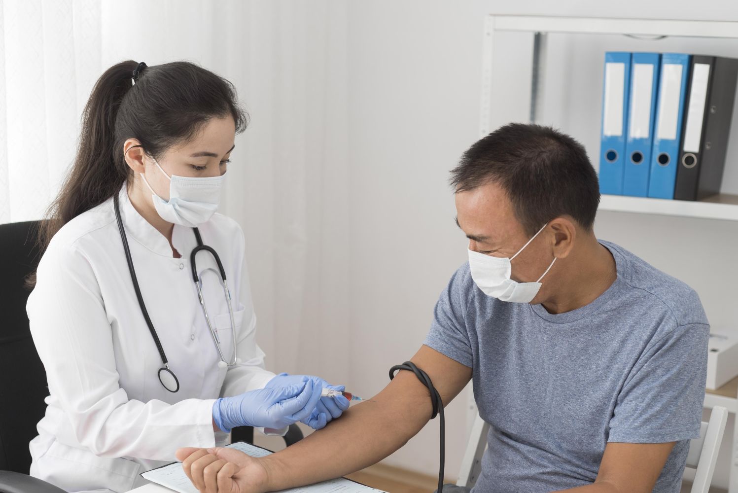 Female doctor in white coat and mask administers a injection to a male patient wearing a mask in a clinic. A stethoscope hangs from her neck.