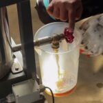Person wearing gloves turns a red valve to release liquid into a translucent beaker on a lab bench area.