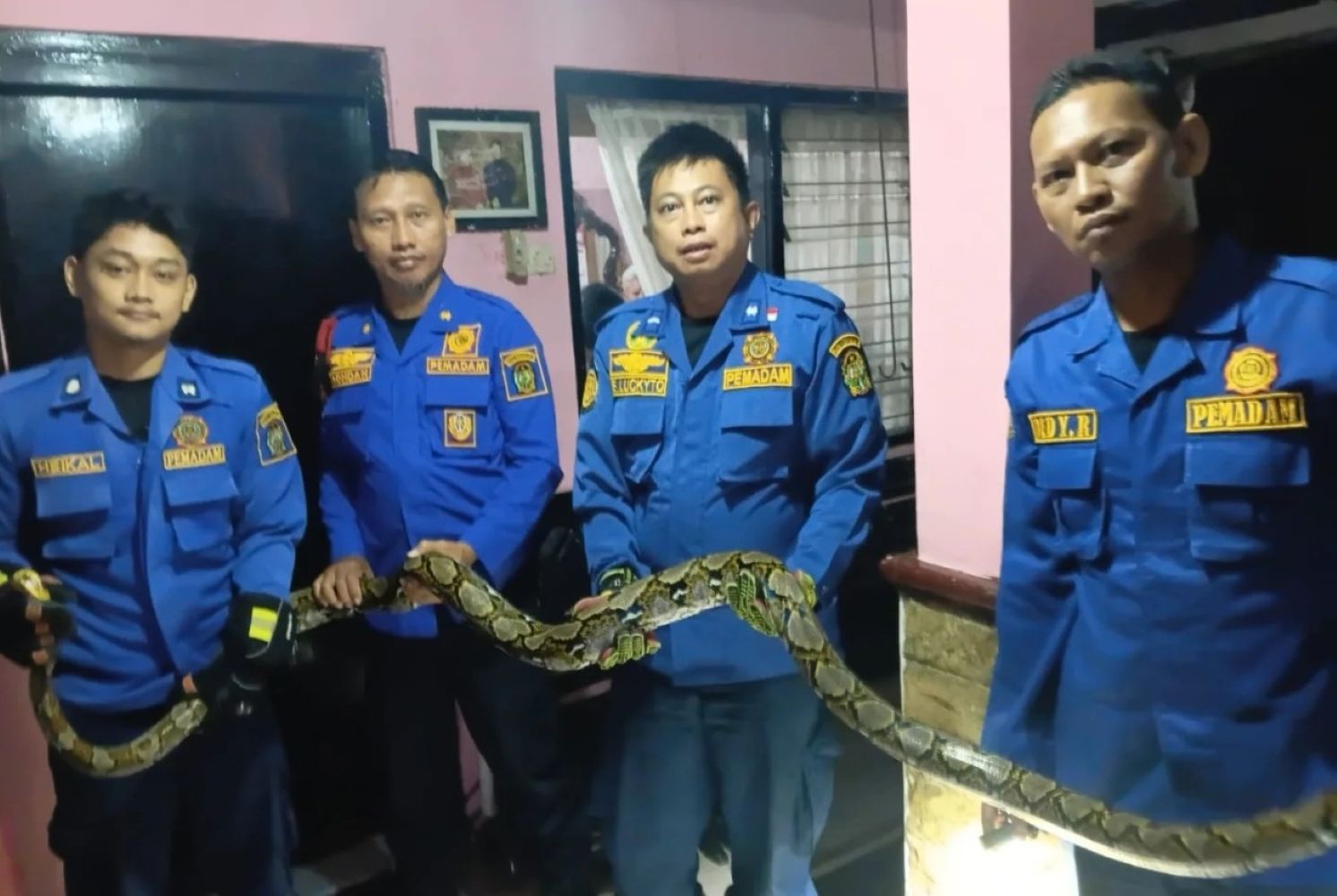 Four men in blue rescue uniforms indoors, holding a long snake together across a railing.