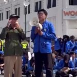 Man in blue jacket speaks into a megaphone while a man in olive green listens during a street protest, with a seated crowd in blue jackets behind them.