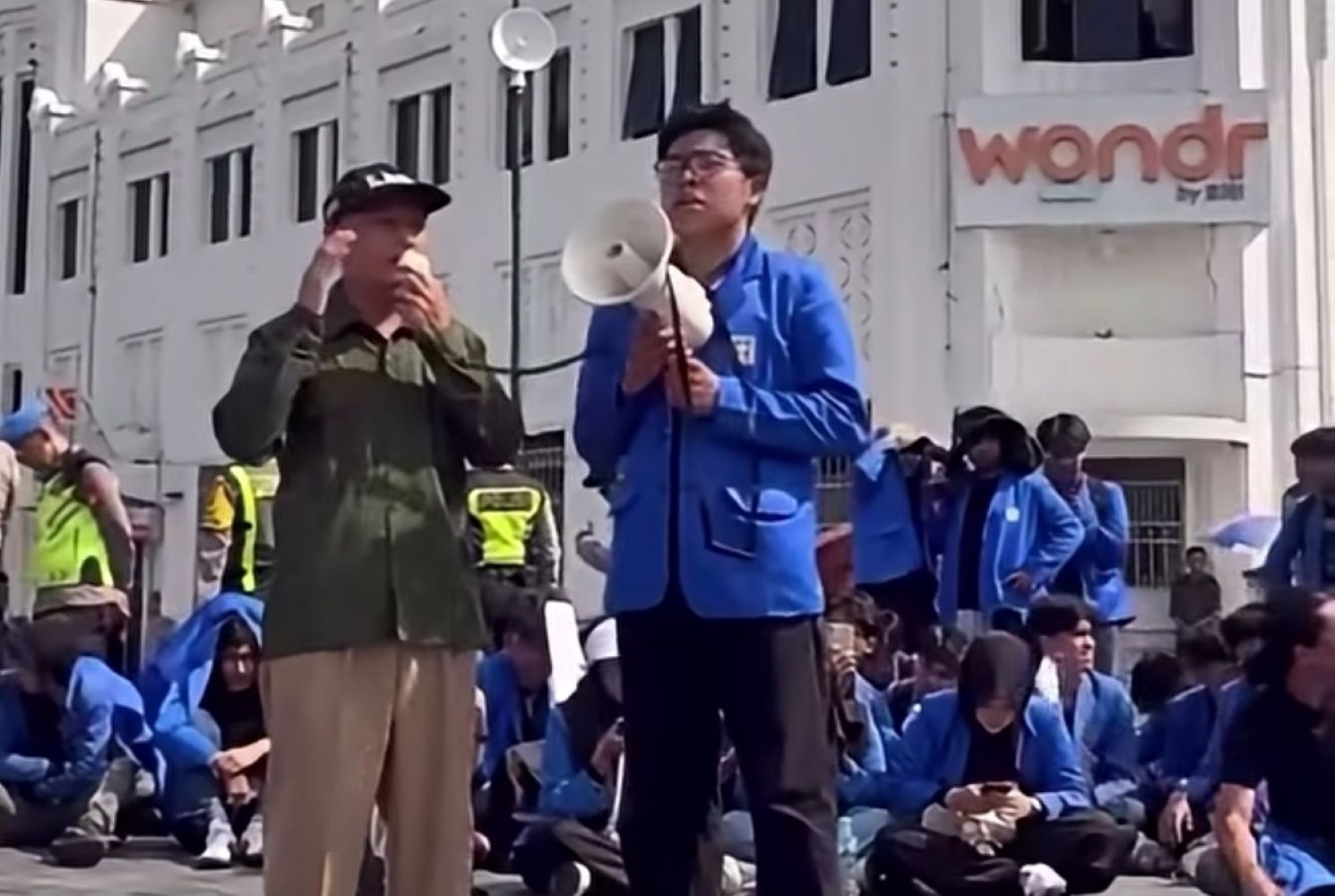 Man in blue jacket speaks into a megaphone while a man in olive green listens during a street protest, with a seated crowd in blue jackets behind them.