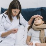 Doctor in a white coat checks a sick girl resting in bed, holding a thermometer and placing a hand on her forehead to feel for fever.
