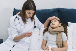 Doctor in a white coat checks a sick girl resting in bed, holding a thermometer and placing a hand on her forehead to feel for fever.