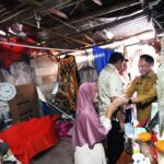 Government official in a khaki uniform shakes hands with a woman wearing a pink headscarf inside a cluttered room filled with boxes, a clock, and various supplies.