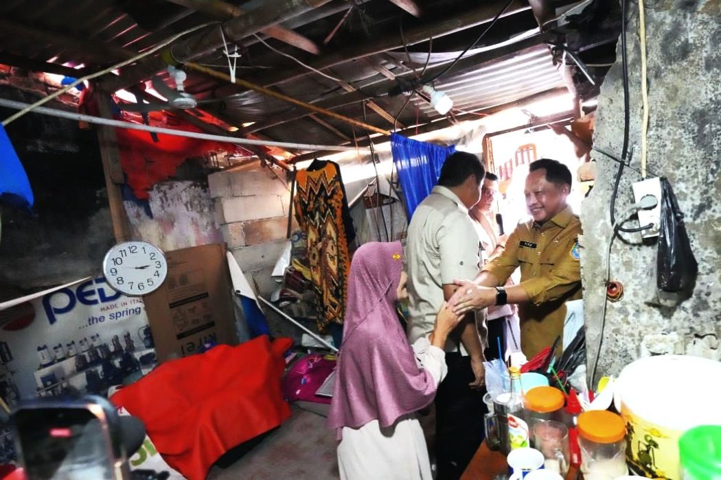 Government official in a khaki uniform shakes hands with a woman wearing a pink headscarf inside a cluttered room filled with boxes, a clock, and various supplies.