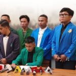 Man in a green blazer signs a document at a press conference while eight colleagues in colorful jackets stand behind him, microphones on the table.