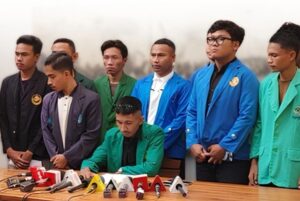 Man in a green blazer signs a document at a press conference while eight colleagues in colorful jackets stand behind him, microphones on the table.