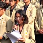 Young woman with glasses speaks into a microphone while reading papers, surrounded by classmates in beige blazers at a school event.