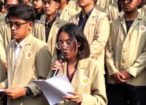 Young woman with glasses speaks into a microphone while reading papers, surrounded by classmates in beige blazers at a school event.