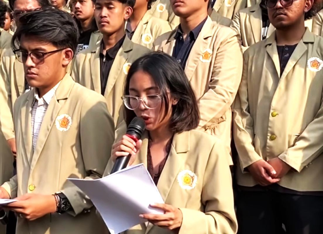 Young woman with glasses speaks into a microphone while reading papers, surrounded by classmates in beige blazers at a school event.