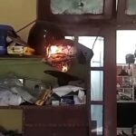 Helmet on a cluttered shelf is burning, with visible flames and smoke in a dim room.