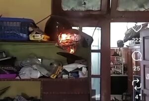 Helmet on a cluttered shelf is burning, with visible flames and smoke in a dim room.