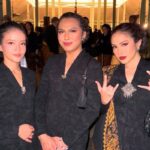 Three women in matching black patterned jackets posing together at an indoor evening event, two with hands clasped in front and the rightmost making a rock-on gesture with a black quilted bag at her side.