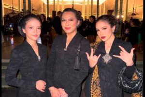 Three women in matching black patterned jackets posing together at an indoor evening event, two with hands clasped in front and the rightmost making a rock-on gesture with a black quilted bag at her side.