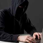 Person in a dark hoodie typing on a laptop, face obscured by the hood in a dim setting.