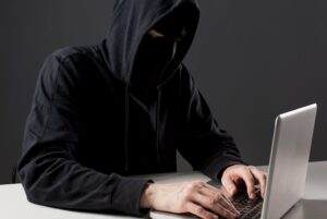 Person in a dark hoodie typing on a laptop, face obscured by the hood in a dim setting.