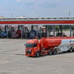 Red fuel tanker truck with a white trailer in the foreground at a Pertamina fuel depot; multiple loading bays and other trucks visible in the background.