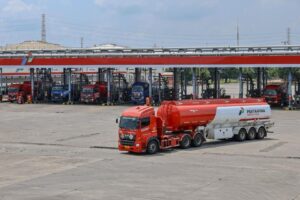 Red fuel tanker truck with a white trailer in the foreground at a Pertamina fuel depot; multiple loading bays and other trucks visible in the background.