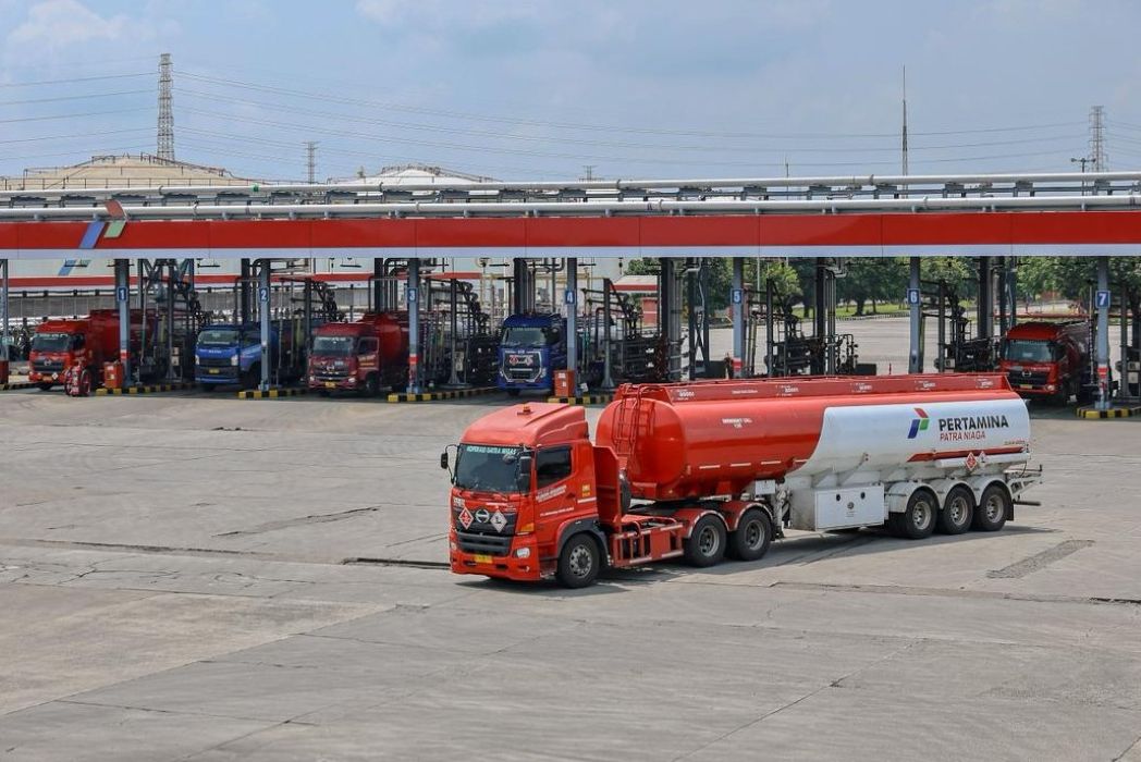 Red fuel tanker truck with a white trailer in the foreground at a Pertamina fuel depot; multiple loading bays and other trucks visible in the background.