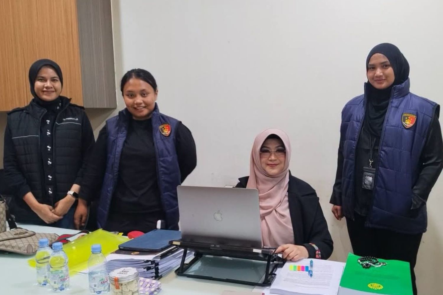 Four women in an office setting: one seated at a desk using a laptop, while three others stand beside her wearing blue padded vests with a crest.