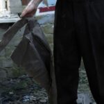 Person in black pants stands among rubble, gripping a torn dark metal sheet; blood on the right hand.