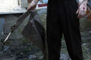 Person in black pants stands among rubble, gripping a torn dark metal sheet; blood on the right hand.
