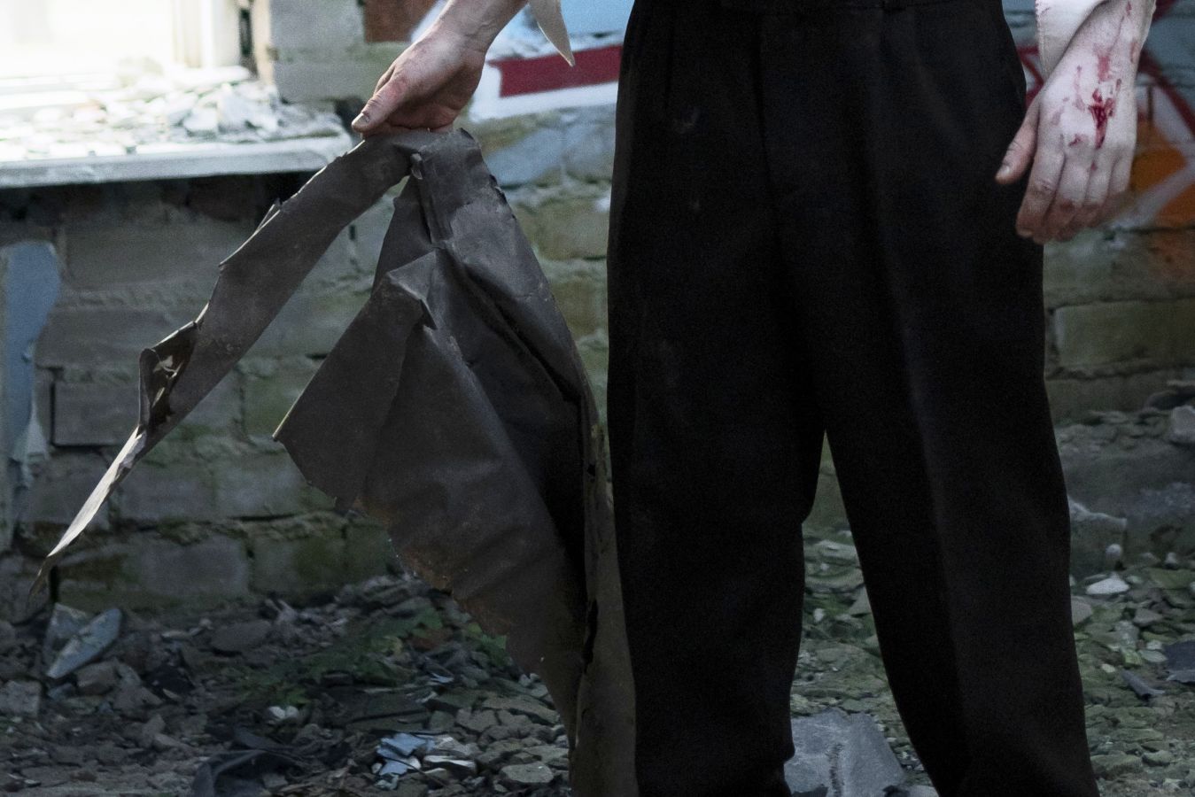 Person in black pants stands among rubble, gripping a torn dark metal sheet; blood on the right hand.