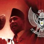 Man in a traditional cap speaks into a microphone, with the Indonesian Garuda emblem in the red background behind him.