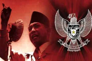 Man in a traditional cap speaks into a microphone, with the Indonesian Garuda emblem in the red background behind him.