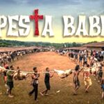 Festival banner reading 'Pesta Babi' above a village square as people carry a pig carcass on a pole in a long line of spectators and participants.