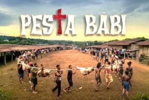 Festival banner reading 'Pesta Babi' above a village square as people carry a pig carcass on a pole in a long line of spectators and participants.