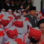 Crowd of people in red-and-white outfits waving Indonesian flags at a public ceremony; a smiling woman stands in the foreground.