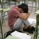 Student sitting on the classroom floor with head tucked into arms, surrounded by desks and scattered papers.