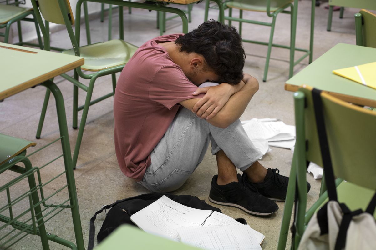 Student sitting on the classroom floor with head tucked into arms, surrounded by desks and scattered papers.