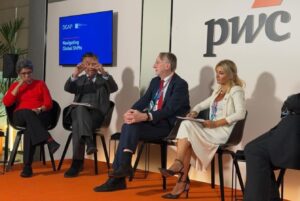 Panel of four professionals seated on stage in a discussion, PwC logo visible behind them and a blue screen reading 'Navigating Global Shifts' on the left.